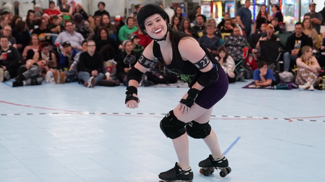 A roller derby skater smiles at the camera while a crowd behind watches on